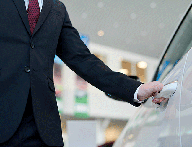 A professional chauffeur in a suit and red tie opening the door of a white luxury car, representing the premium chauffeured car rental service in Istanbul.